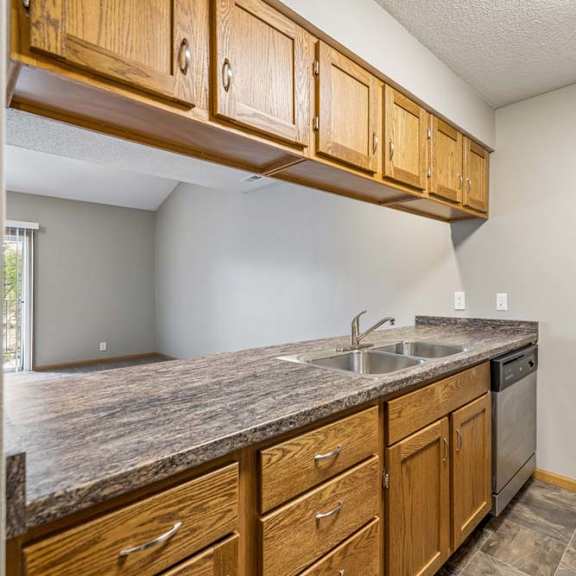 Thumbnail 14 of 26 Open apartment kitchen with wood-tone cabinets, laminate countertops, a double-basin sink, dishwasher, overhead storage, and a breakfast bar overlooking the living room.