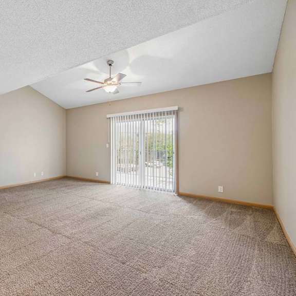 Thumbnail 18 of 26 Spacious, empty apartment living room with vaulted ceilings, neutral carpet, a ceiling fan, and a large sliding glass door with vertical blinds that fills the space with natural light.