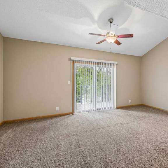Thumbnail 17 of 26 Bright, empty apartment living room with vaulted ceilings, neutral carpet, ceiling fan, and a large sliding glass door with vertical blinds that lets in natural light.