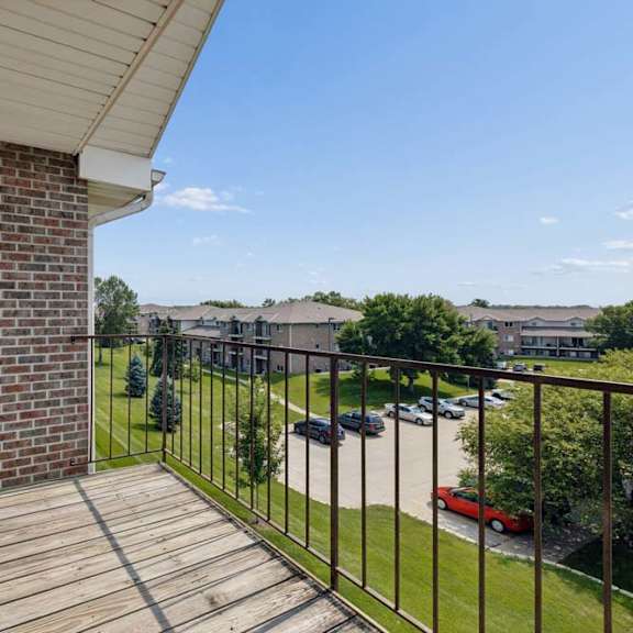 Thumbnail 19 of 21 Upper-level balcony with wood decking and metal railing overlooking landscaped community grounds and nearby apartment buildings.
