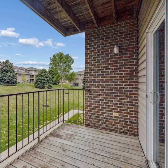 Thumbnail 31 of 31 Covered apartment balcony with wood decking, a brick accent wall, metal railing, and views of the landscaped grounds and neighboring apartment buildings.
