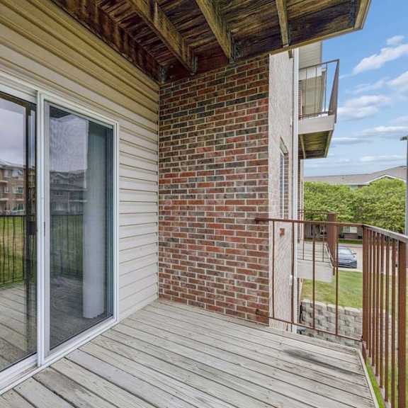 Thumbnail 30 of 31 Covered apartment balcony with wood decking, a sliding glass door, brick accent wall, metal railing, and views of the surrounding apartment community and green space.