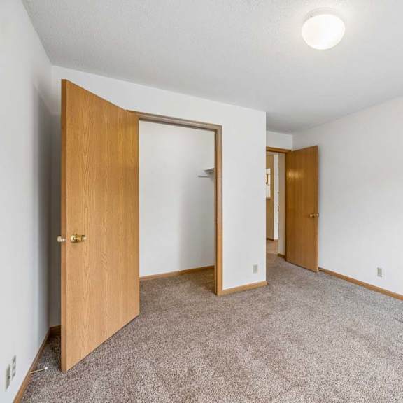 Thumbnail 25 of 31 Bright, empty apartment bedroom with neutral carpet, white walls, wood-trimmed doors, and an open closet area leading to the adjoining hallway.