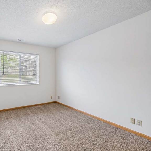 Thumbnail 21 of 31 Bright, empty apartment bedroom with neutral carpet, white walls, wood trim, and a single window with blinds providing natural light.
