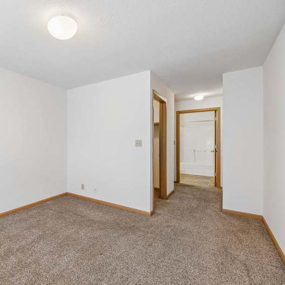 Thumbnail 24 of 31 Bright, empty apartment bedroom with neutral carpet, white walls, and an open layout leading to the hallway and adjoining bathroom.