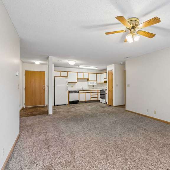 Thumbnail 19 of 31 Spacious apartment living room with neutral carpet, a ceiling fan, and an open layout leading to the kitchen with a breakfast bar, wood-tone cabinets, and white appliances.