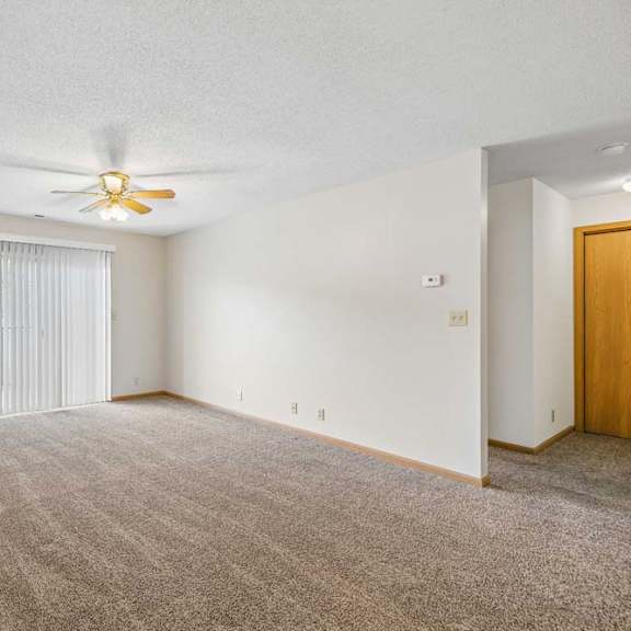 Thumbnail 20 of 31 Spacious apartment living room with neutral carpet, a ceiling fan, vertical blinds over a sliding glass door, and an open layout leading to the hallway.