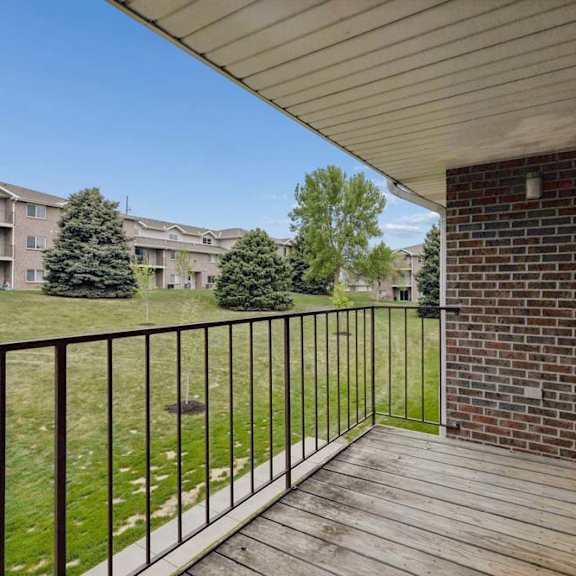 Thumbnail 18 of 18 Covered apartment balcony with wood decking, metal railings, and views of landscaped green space and neighboring apartment buildings.