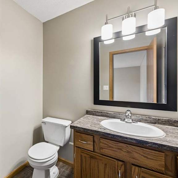 Thumbnail 16 of 18 Apartment bathroom with a single-sink vanity, wood cabinetry, a granite-look countertop, a framed mirror with modern lighting, a tub-shower combination, and an adjacent walk-in closet.
