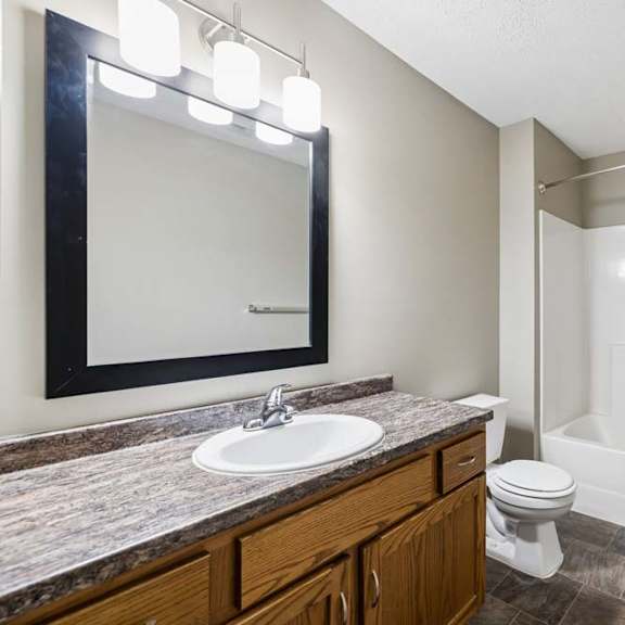 Thumbnail 15 of 18 Apartment bathroom with a single-sink vanity, wood cabinetry, a granite-look countertop, a framed mirror with modern lighting, and a tub-shower combination with a white surround.