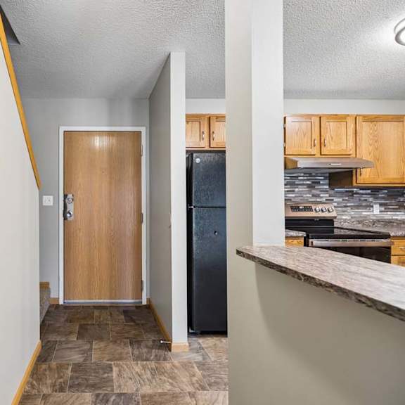 Thumbnail 6 of 18 Apartment entryway with tile flooring, a wood front door, and a breakfast bar leading into the kitchen with wood cabinetry, black appliances, and a tile backsplash.