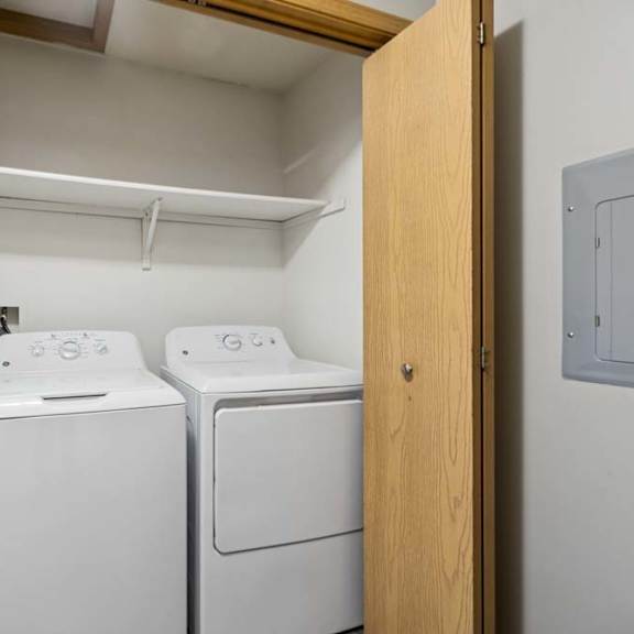 Thumbnail 17 of 18 Apartment laundry closet with side-by-side washer and dryer, built-in shelf above, and wood bifold doors for convenient in-home laundry access.