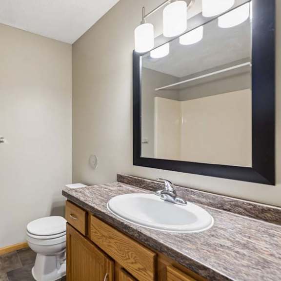 Thumbnail 26 of 28 Apartment bathroom with a single-sink vanity, wood cabinetry, a granite-look countertop, a framed mirror with modern lighting, and a tub-shower combination with a white surround.