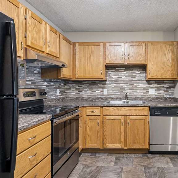 Thumbnail 17 of 28 Apartment kitchen with wood cabinetry, a tile backsplash, laminate countertops, black and stainless steel appliances, and a functional layout adjacent to the entryway.
