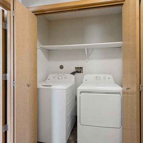Thumbnail 27 of 28 Apartment laundry closet with side-by-side washer and dryer, a built-in shelf above, and wood bifold doors for convenient in-home laundry access.