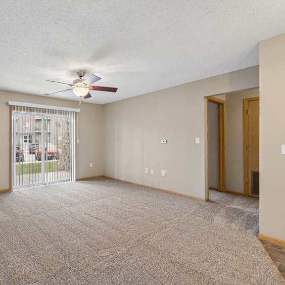Thumbnail 20 of 28 Spacious apartment living room with carpeted flooring, a ceiling fan, and a large sliding glass door with vertical blinds providing natural light and access to a private patio or balcony.