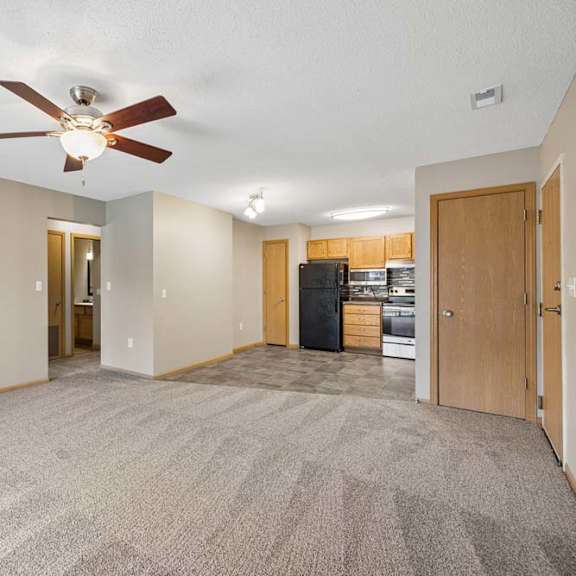 Thumbnail 18 of 28 Spacious open-concept apartment living room with carpeted flooring, a ceiling fan, and a view into the kitchen featuring wood cabinetry, black appliances, and an adjacent dining area.