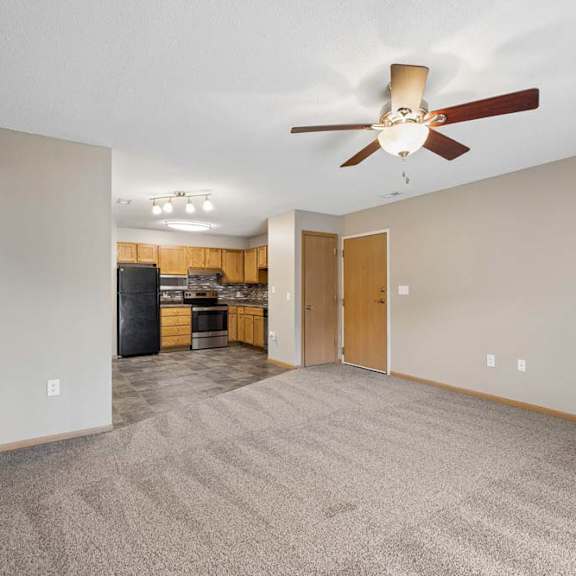 Thumbnail 19 of 28 Spacious open-concept apartment living room with carpeted flooring, a ceiling fan, and a view into the kitchen featuring wood cabinetry, black appliances, and an adjacent dining area.