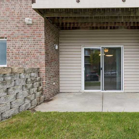 Thumbnail 28 of 28 Covered ground-floor apartment patio with a concrete surface, a large sliding glass door, brick and siding exterior, and direct access to a landscaped lawn.