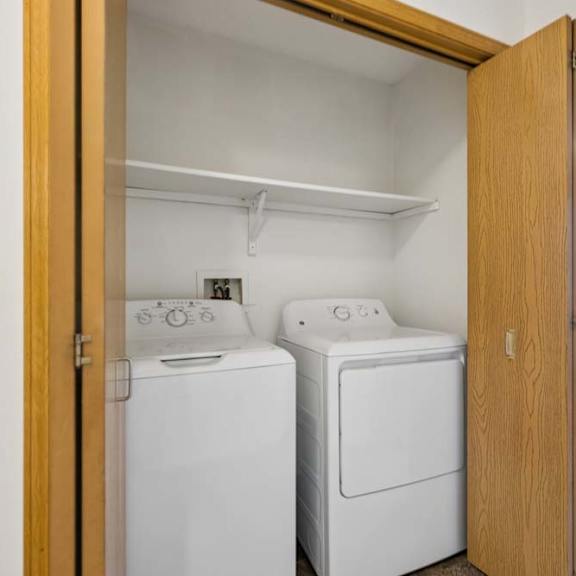 Thumbnail 21 of 23 Apartment laundry closet with side-by-side washer and dryer, a built-in shelf above, and wood bifold doors for convenient in-home laundry access.