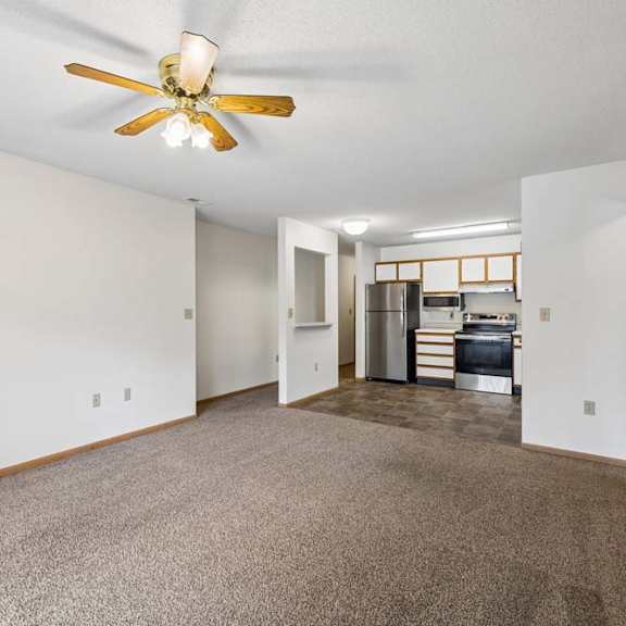 Thumbnail 13 of 23 Spacious open-concept apartment living room with carpeted flooring, a ceiling fan, and a view into the kitchen featuring white and wood-tone cabinetry, stainless steel and black appliances, and a functional open layout.