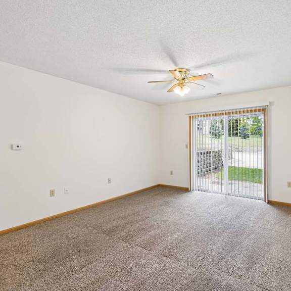 Thumbnail 15 of 23 Spacious apartment living room with carpeted flooring, a ceiling fan, and a large sliding glass door with vertical blinds providing natural light and access to a private patio or balcony.