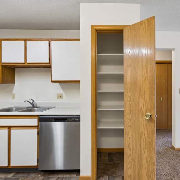 Thumbnail 14 of 23 Apartment pantry with built-in shelving and a wood-finish door, located just off the kitchen with white and wood-tone cabinetry and adjacent open living space.