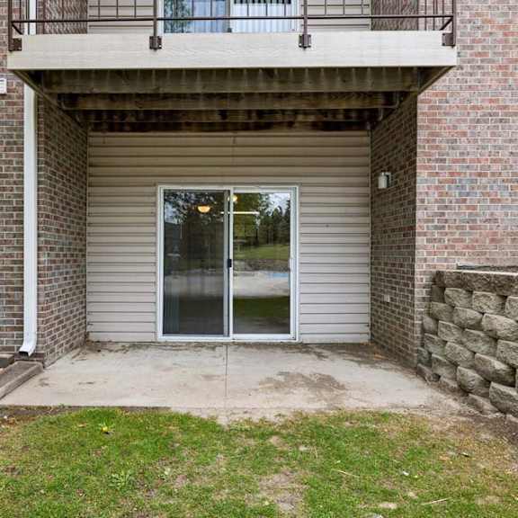 Thumbnail 23 of 23 Covered ground-floor apartment patio with a concrete surface, a large sliding glass door, brick and siding exterior, and direct access to a landscaped lawn.