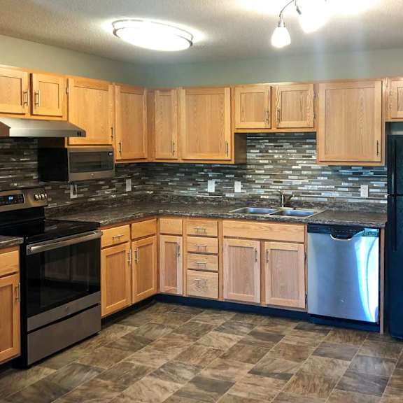 Thumbnail 2 of 6 A kitchen with wooden cabinets, a black refrigerator, stainless steel appliances, a tile backsplash, and granite countertops.