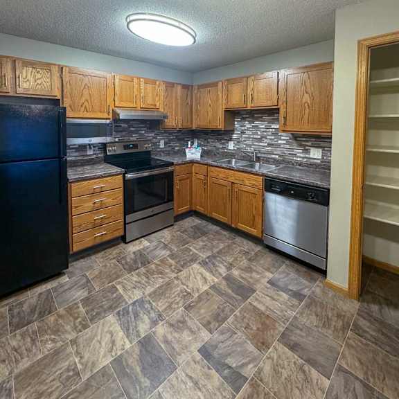 Thumbnail 3 of 9 A kitchen with a black refrigerator, wooden cabinets, stainless steel appliances, a tile backsplash, and granite countertop