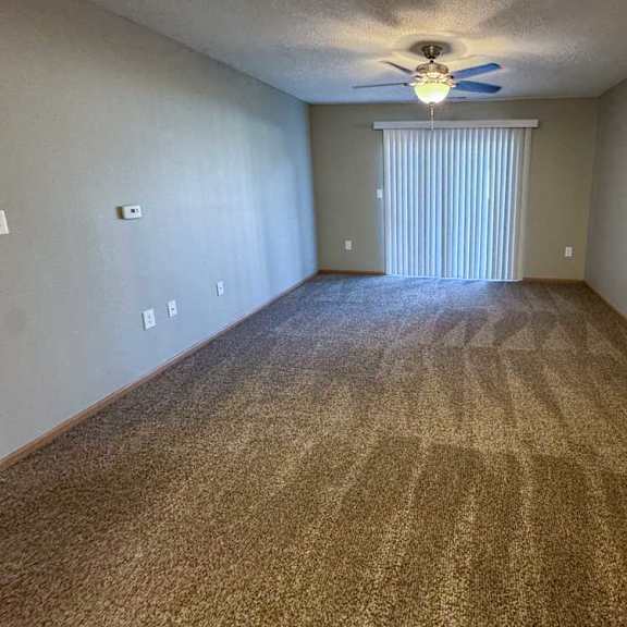 Thumbnail 4 of 9 A living room with a carpeted floor, ceiling fan, and a sliding glass door with vertical blinds.