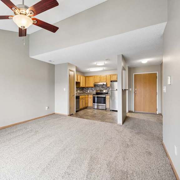 Thumbnail 7 of 20 A room with a ceiling fan and carpeted floor and a kitchen in the background.