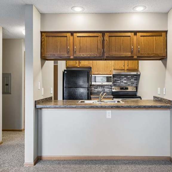 Thumbnail 22 of 29 Open apartment kitchen with wood cabinetry, black appliances, a breakfast bar, and a pass-through layout connecting the kitchen to the living area.