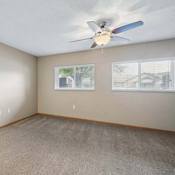 Thumbnail 25 of 29 Spacious apartment bedroom with carpeted flooring, a ceiling fan, and two windows with blinds providing natural light and neighborhood views.