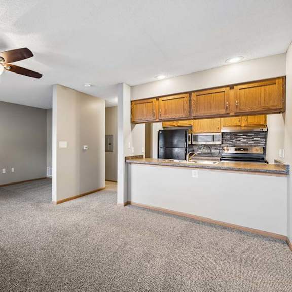 Thumbnail 21 of 29 Open-concept apartment living room with carpeted flooring, a ceiling fan, and a breakfast bar overlooking the kitchen with wood cabinetry and black appliances