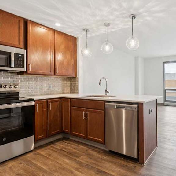 Thumbnail 7 of 16 A kitchen with wooden cabinets and a stainless steel dishwasher, in front of a living room with a large window.