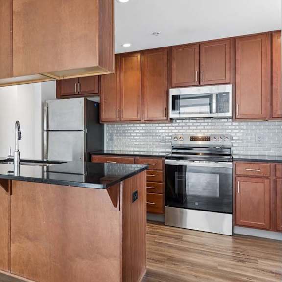 Thumbnail 3 of 19 Apartment kitchen with wood cabinetry, black countertops, stainless steel appliances, tile backsplash, and visible laundry area.