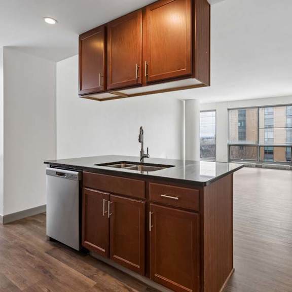 Thumbnail 2 of 19 Bright apartment kitchen with wood cabinetry, dark countertops, stainless dishwasher, and open living area with large windows.