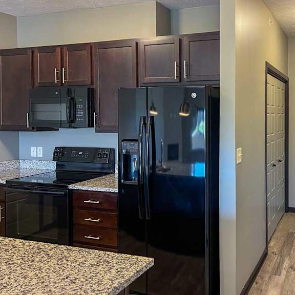 Thumbnail 26 of 35 a kitchen with dark wooden cabinets, granite countertops, and black appliances in front of a hallway to the attached garage