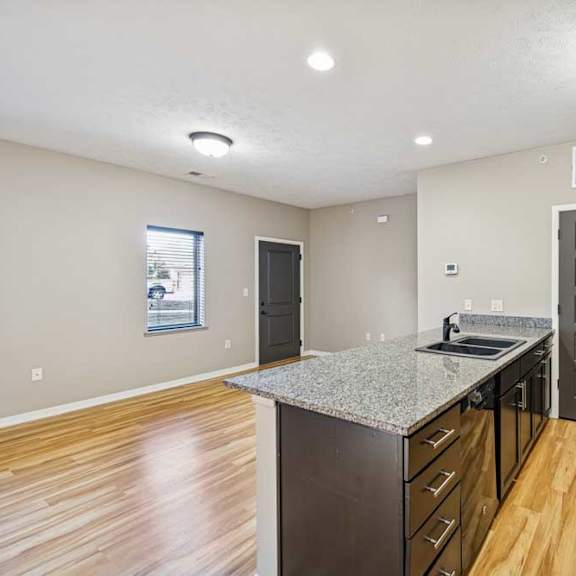 Thumbnail 13 of 18 A kitchen with grey granite countertops, black appliances, and a large kitchen island. There is a large living room with wooden floors throughout. There is a window next to the front door in the living room.