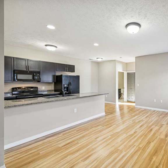 Thumbnail 15 of 18 A large living room with wooden floors and a large kitchen island. Black appliances are in the kitchen. There is a hallway in the background.