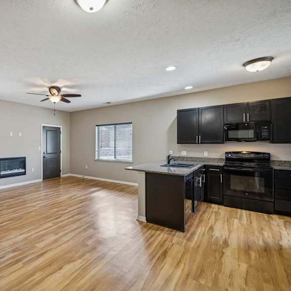 Thumbnail 5 of 22 A spacious living room with hardwood-style flooring and an electric fireplace, open to a kitchen with dark wooden cabinets.