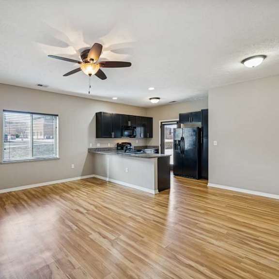 Thumbnail 6 of 22 A spacious living room with hardwood-style flooring and large windows, open to a kitchen with dark wooden cabinets.