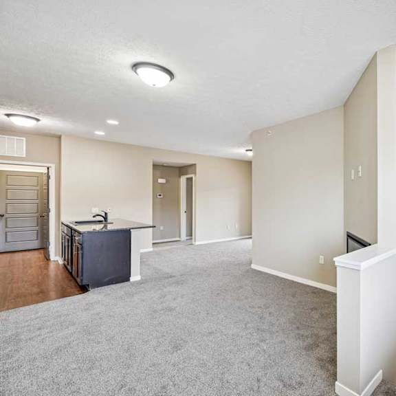 Thumbnail 7 of 25 Open-concept apartment living space with neutral carpeting, soft beige walls, and a half wall that opens to a stairwell below.
