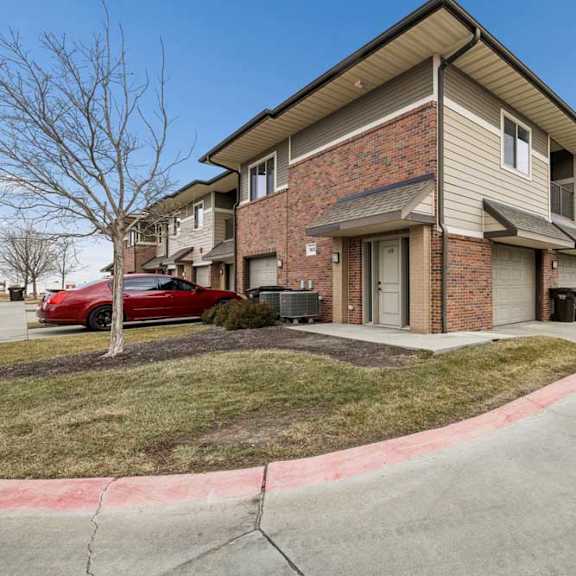 Thumbnail 2 of 25 Exterior view of a two-story apartment building with beige siding, brick accents, and a garage entrance along a curved driveway.