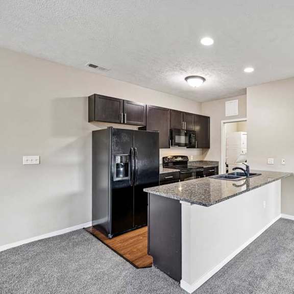 Thumbnail 11 of 25 Open apartment kitchen featuring dark wood cabinets, black appliances, a granite island with sink, and carpeted living space.