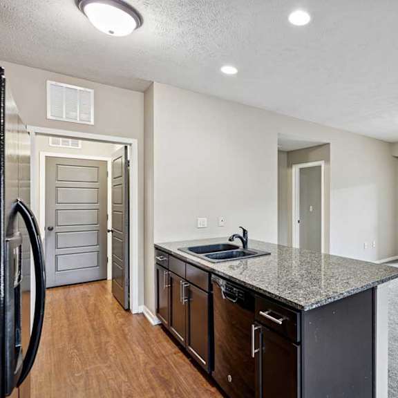 Thumbnail 6 of 25 Open-concept apartment featuring a kitchen with dark wood cabinets and granite island, wood-style flooring, and a carpeted living area with sliding doors to a balcony.