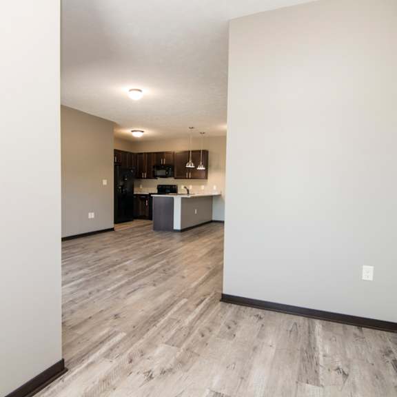 Thumbnail 10 of 14 A room with a kitchen in the background and a wooden floor.