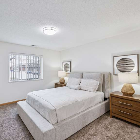 Thumbnail 29 of 56 An apartment bedroom with a large bed, a nightstand, and a window with vertical blinds.