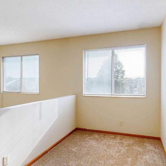 a carpeted loft bedroom with a view open to below, two bright windows, and a ceiling fan.